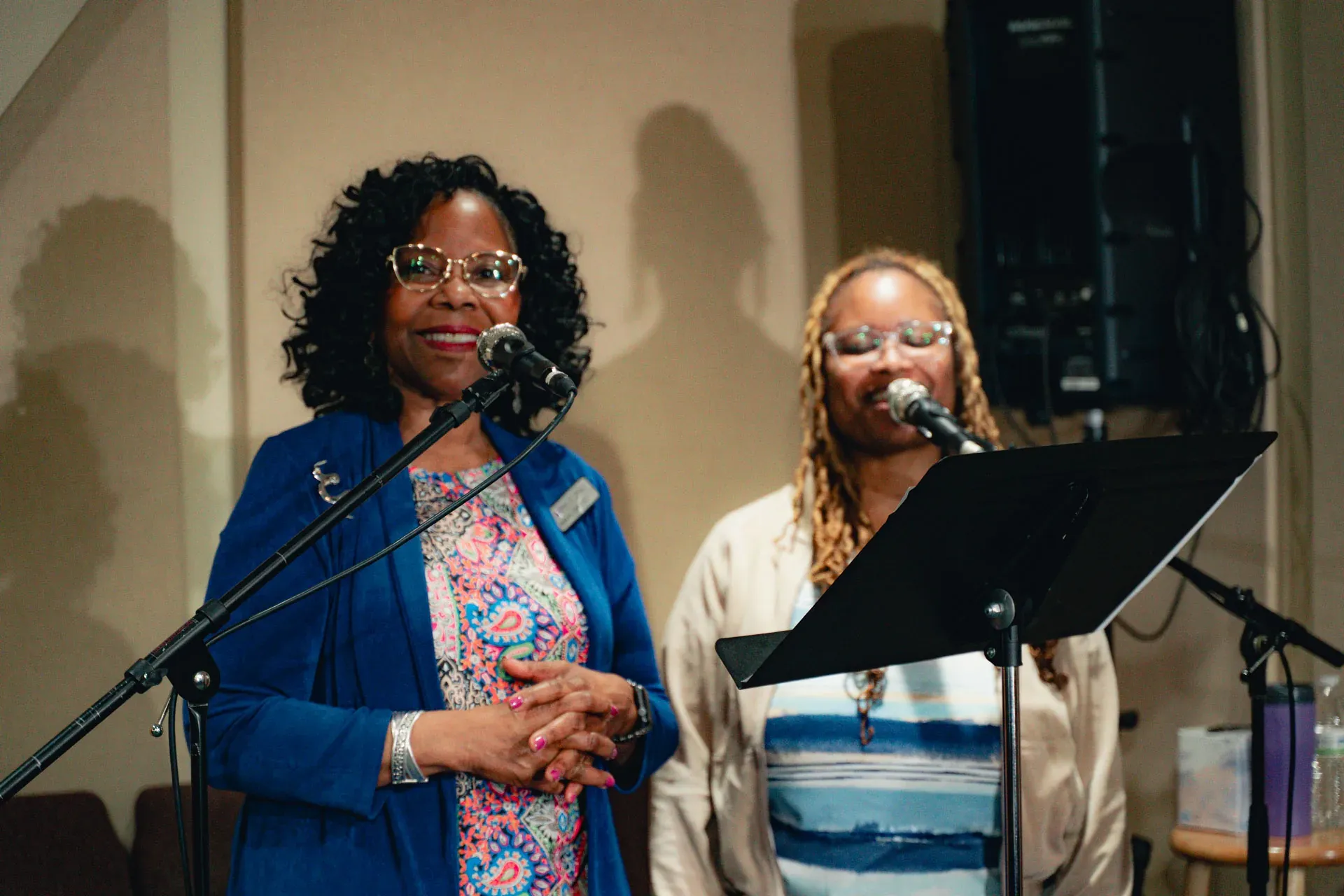 Two women stand at microphones at the front of the sanctuary giving an announcement; one wears a paisley print top with a name badge, the other a striped wrap.