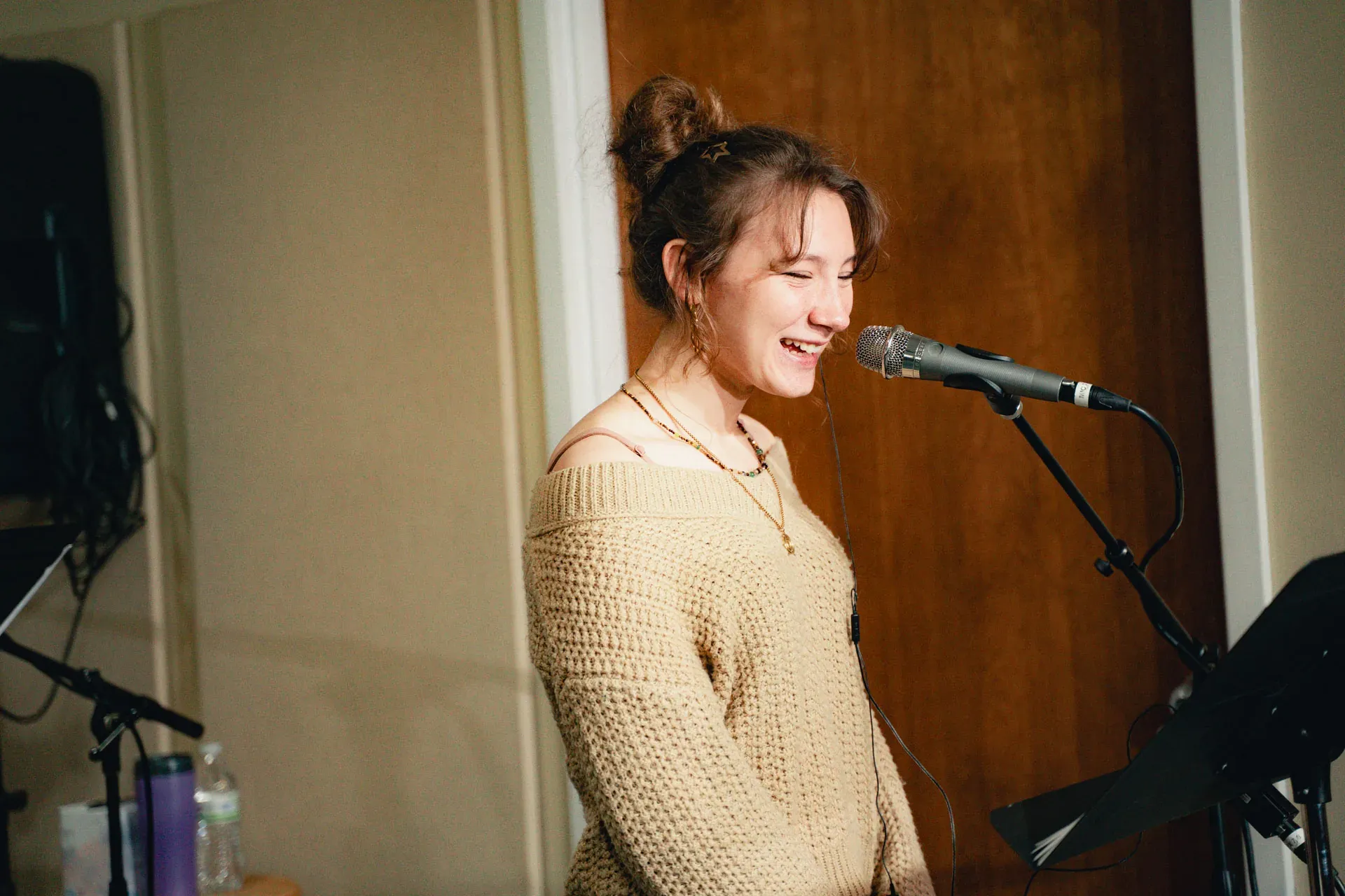 A young woman with auburn hair pulled into a loose top-knot, wearing an oatmeal-colored chunky knit sweater, sings into a microphone with eyes closed, mid-lyric, smiling broadly.