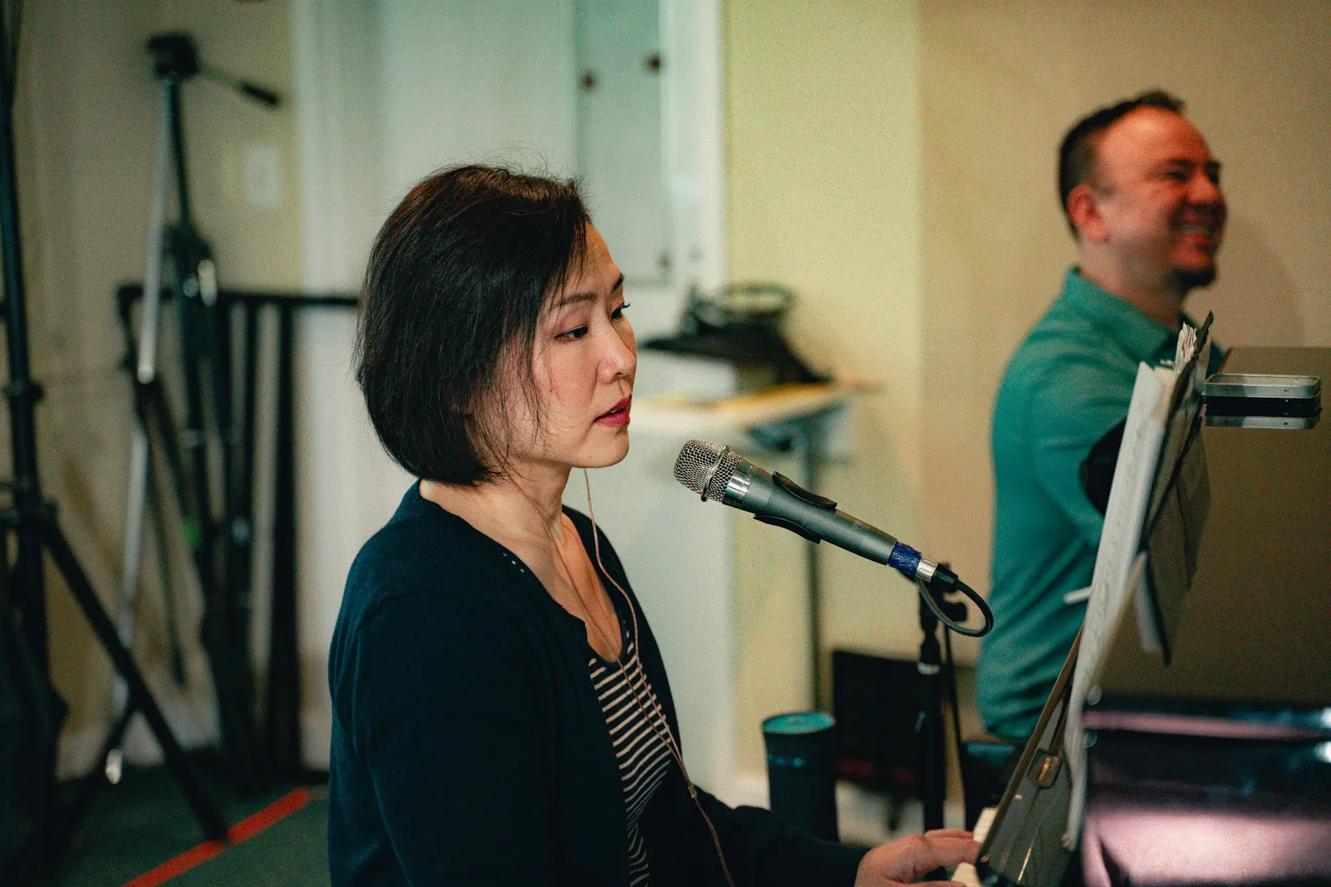 A keyboardist with a chin-length bob, in a dark cardigan over a striped top, sings into a microphone at her keyboard with eyes downcast on the keys; the drummer is just visible in the background, smiling.