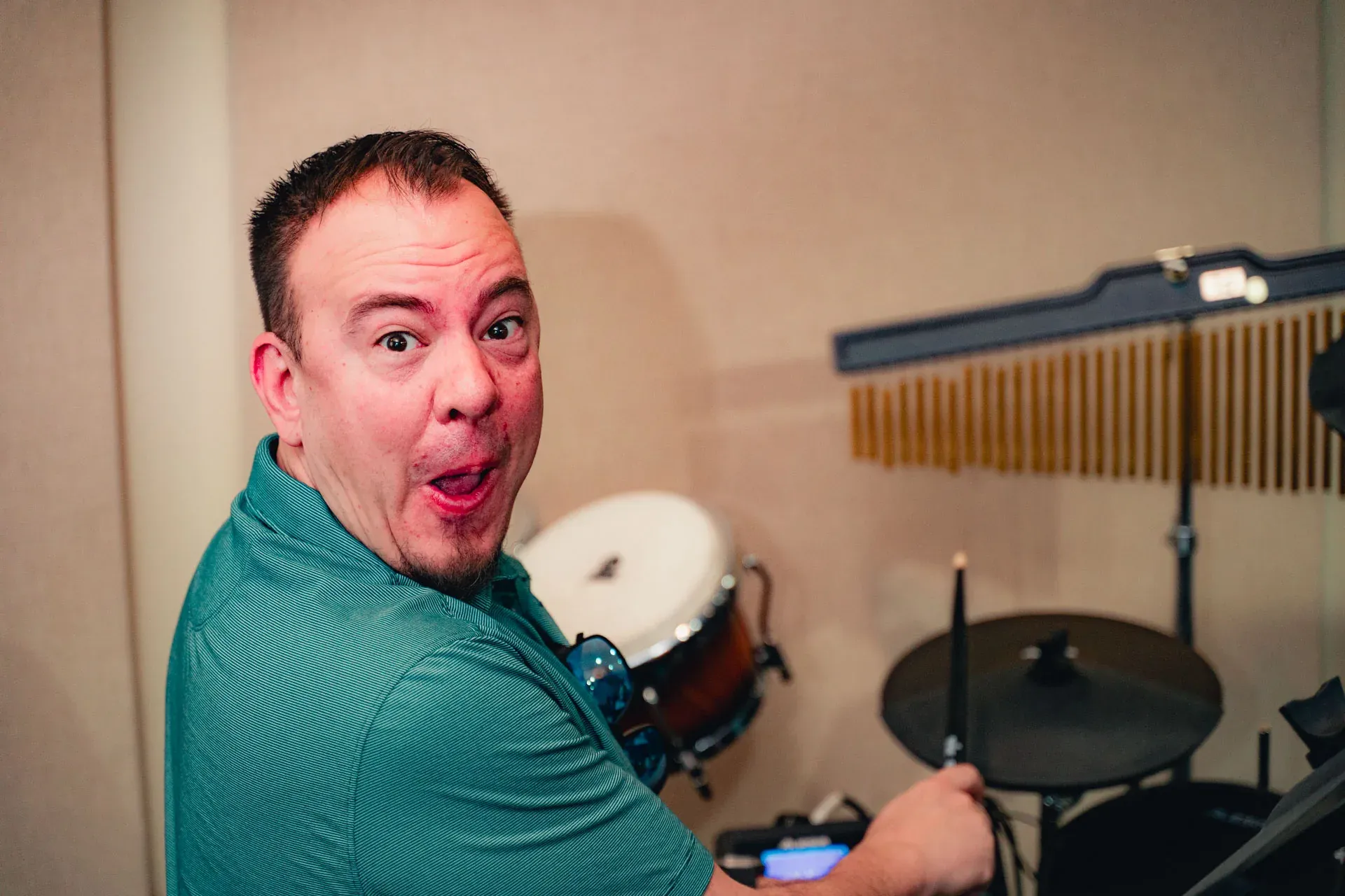 Close portrait of the drummer in a teal polo, turned toward the camera with an open-mouthed expression of mock surprise; a conga drum and wind chimes sit beside the kit.