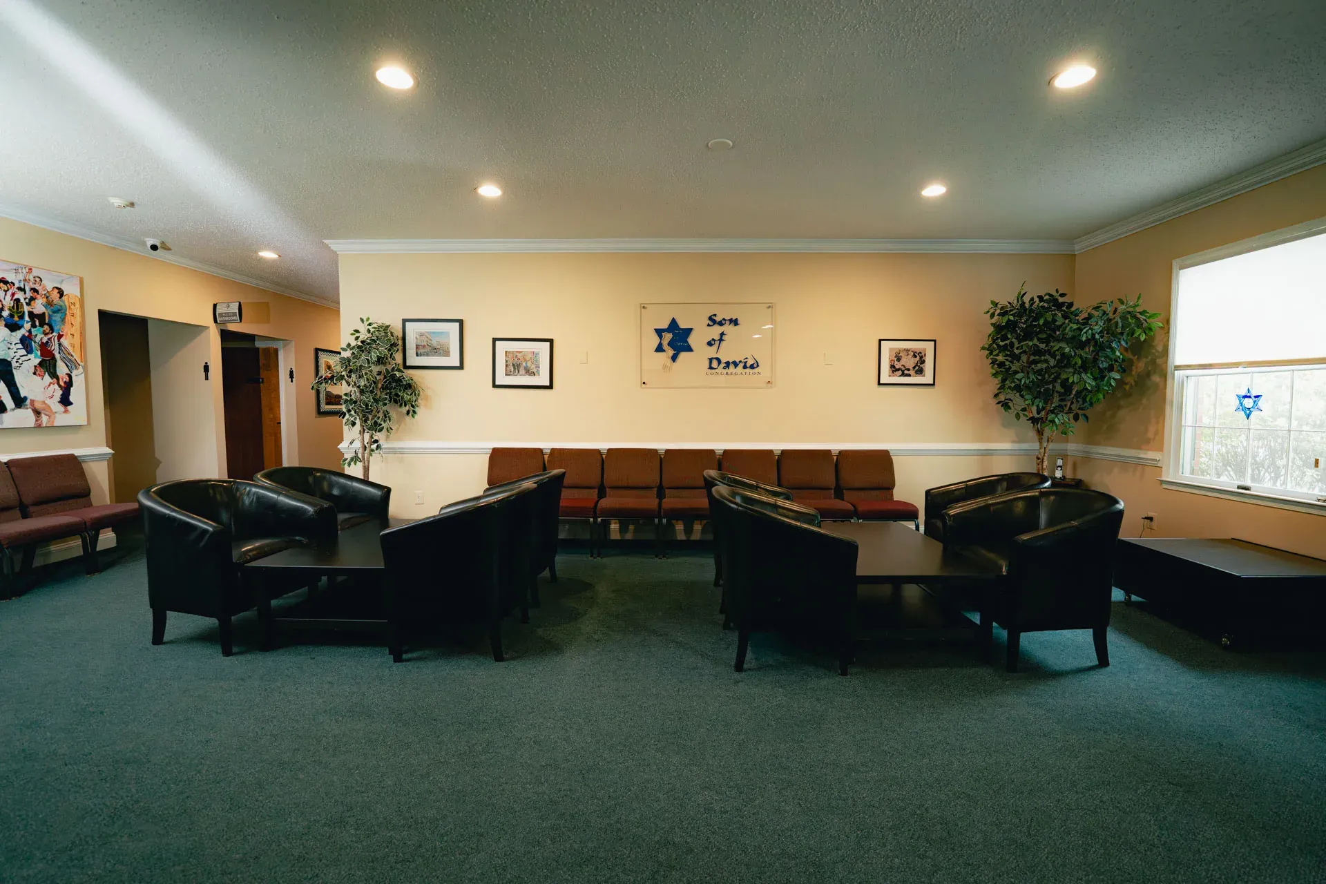 The Son of David Congregation lobby: cream walls, dark green carpet, two clusters of black leather chairs around dark coffee tables, and a row of red upholstered chairs along the back wall under the SODC sign with a blue Star of David.