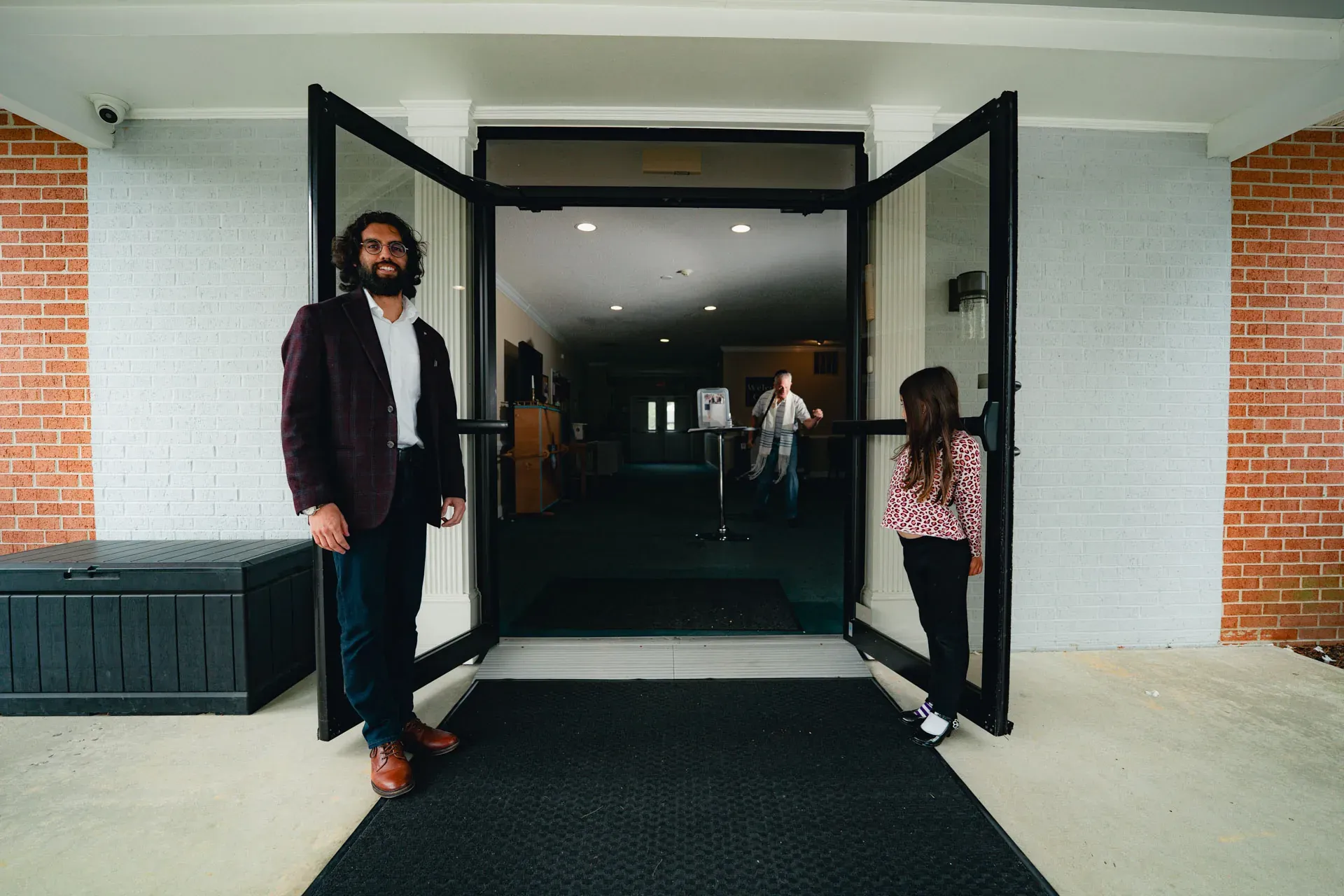 A man holds open one of the church's tall front double doors; a young girl in a pink top stands at the other door, looking inside toward the lobby.