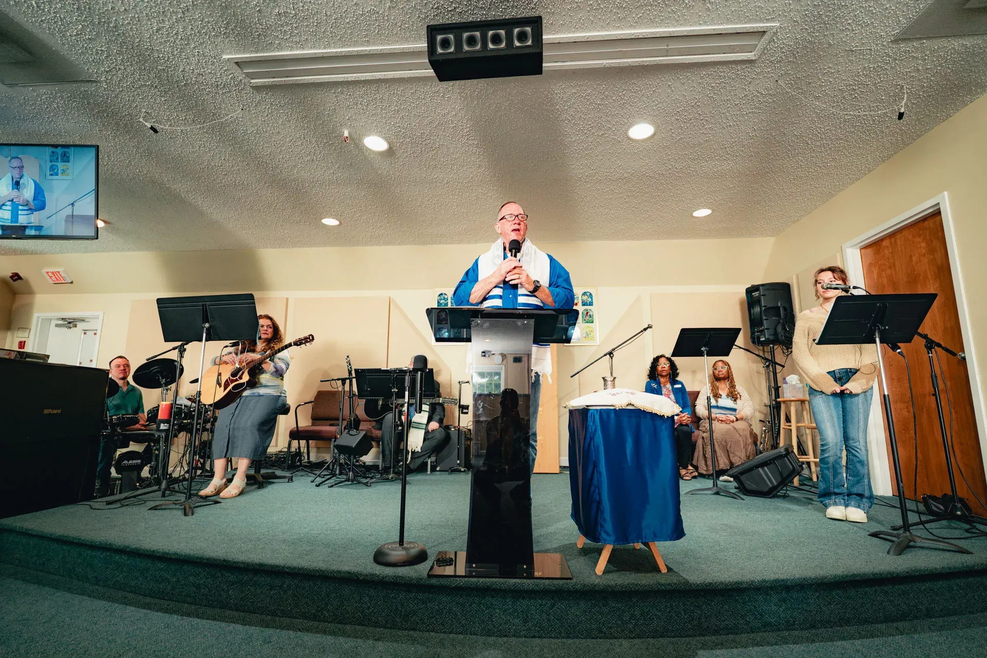 The rabbi at a wood podium in a tallit, speaking into a handheld microphone, with the worship band of guitarists, keyboardist and singers standing behind him on the bimah.