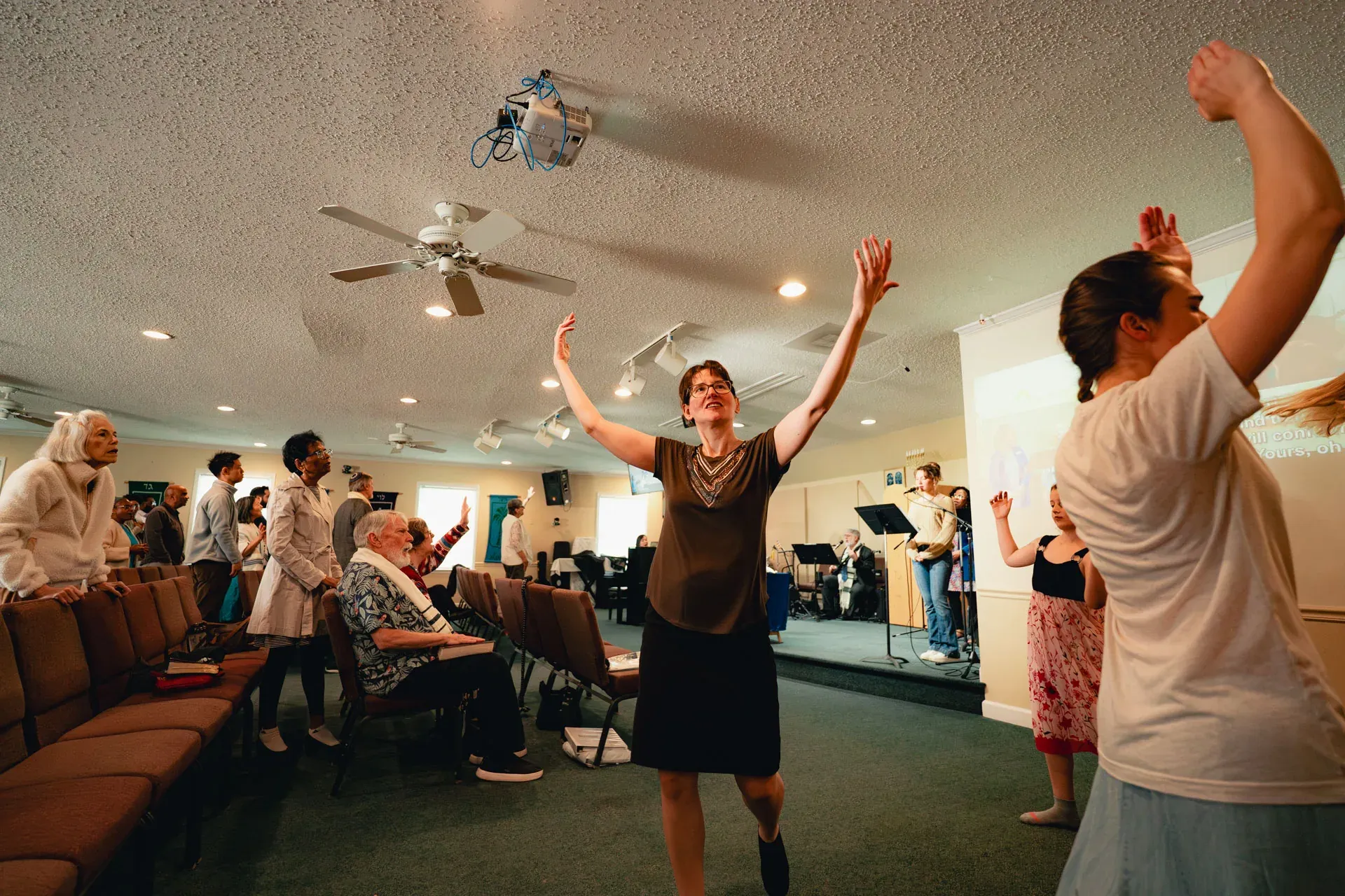 Congregants standing in the aisle with arms raised in worship; the band plays in the background under a ceiling fan.