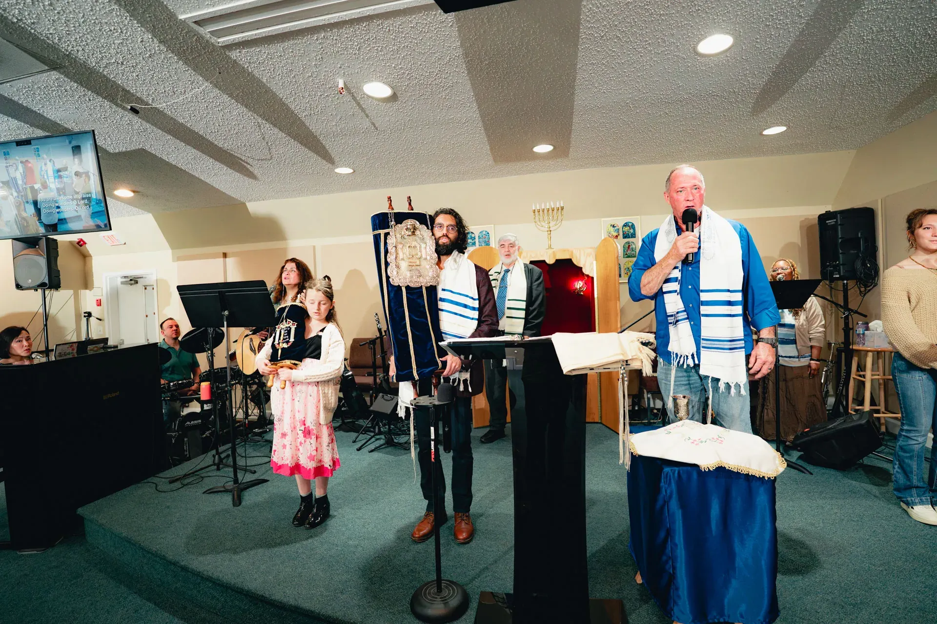 Torah service in progress: the rabbi at the podium in a tallit speaks into a microphone, while a bearded man beside him in a tallit holds the dressed Torah scroll upright. A young girl in a pink dress stands holding a small child-size Torah.