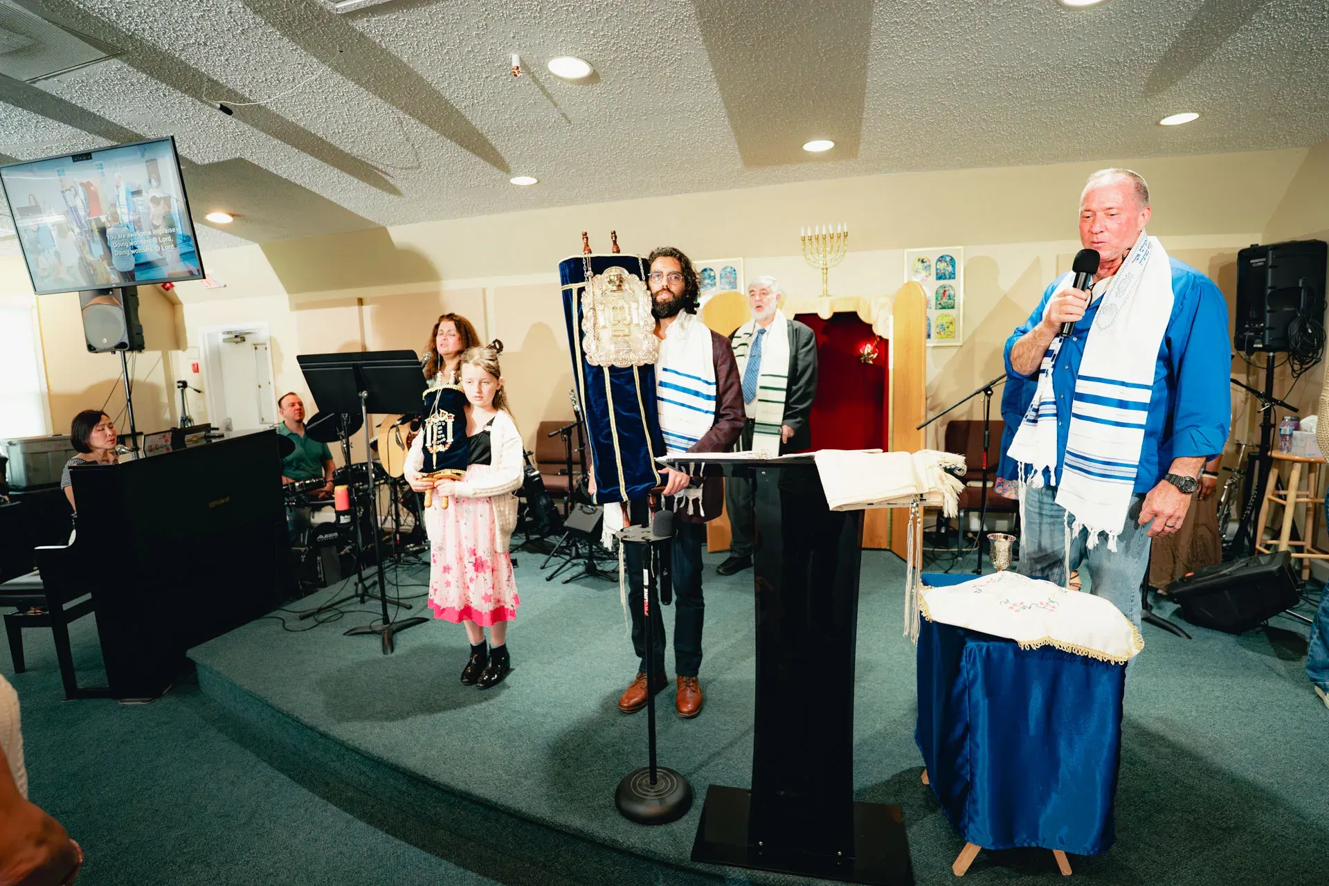 A wide angle of the Torah service: pianist at the keyboard at far left, a young girl in a pink dress with a child-size Torah, a bearded man in a tallit holding the full Torah scroll, the rabbi at the podium speaking. The Aron Kodesh and seven-branch menorah are visible at the rear.