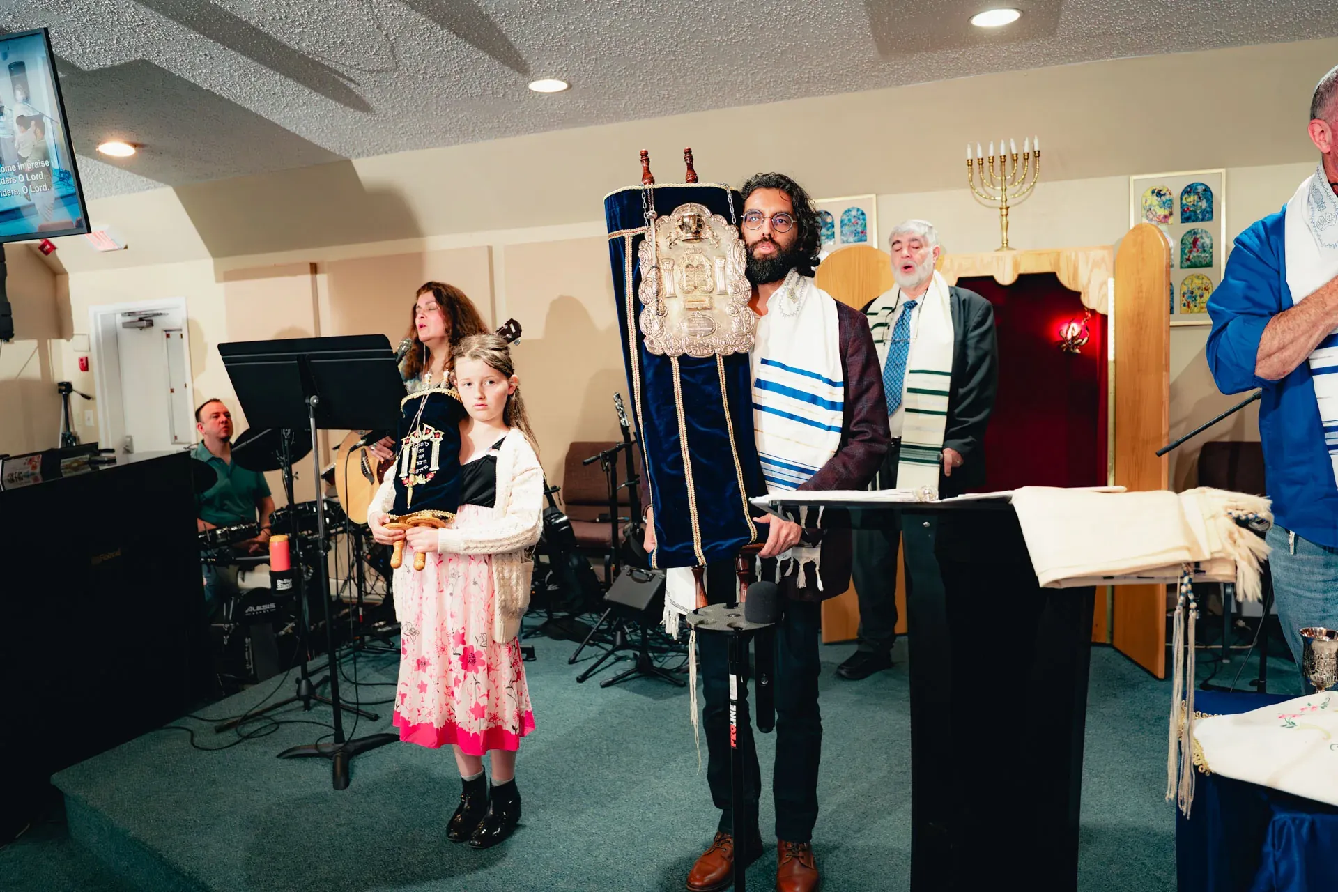 The Aron Kodesh stands open at the front of the sanctuary, its red velvet interior visible. A bearded man in a tallit holds a Torah dressed in royal-blue velvet with a silver breastplate; a young girl in a pink floral dress stands beside him holding a small Torah.