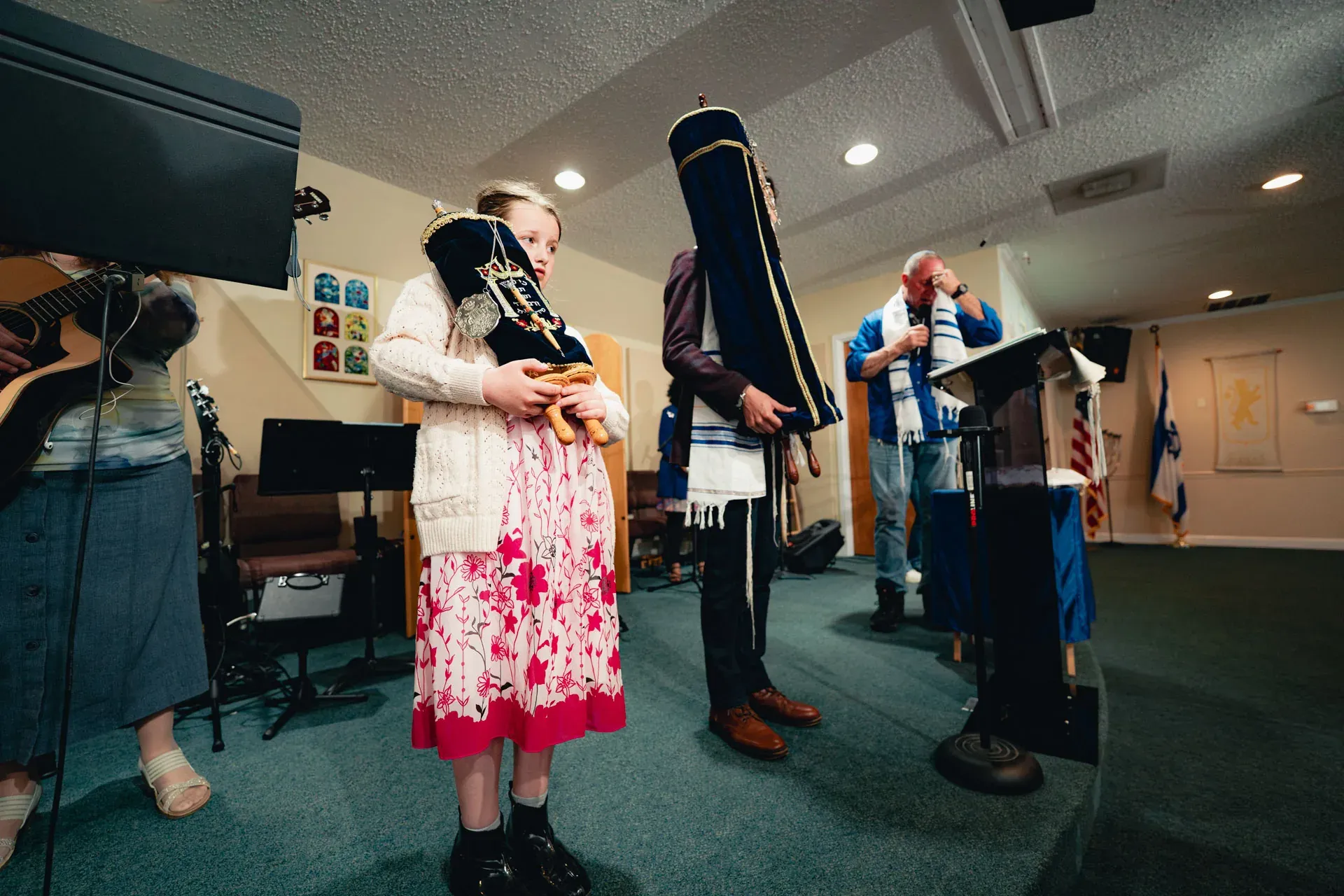 A young blond girl in a pink floral dress and white cardigan holds a small dressed Torah scroll close to her chest, looking down at it. Behind her, a man in a tallit holds the full-size Torah in its blue velvet mantle.
