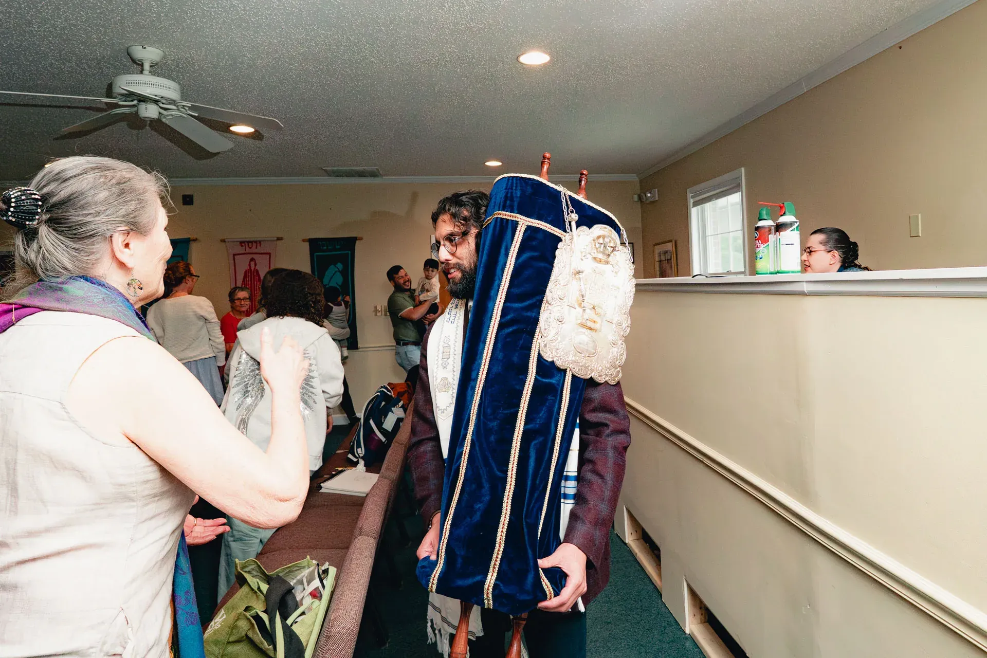 The Torah procession through the back of the sanctuary: a bearded man carries the dressed Torah scroll over his shoulder; an older woman in the foreground reaches toward it with the corner of her tallit.