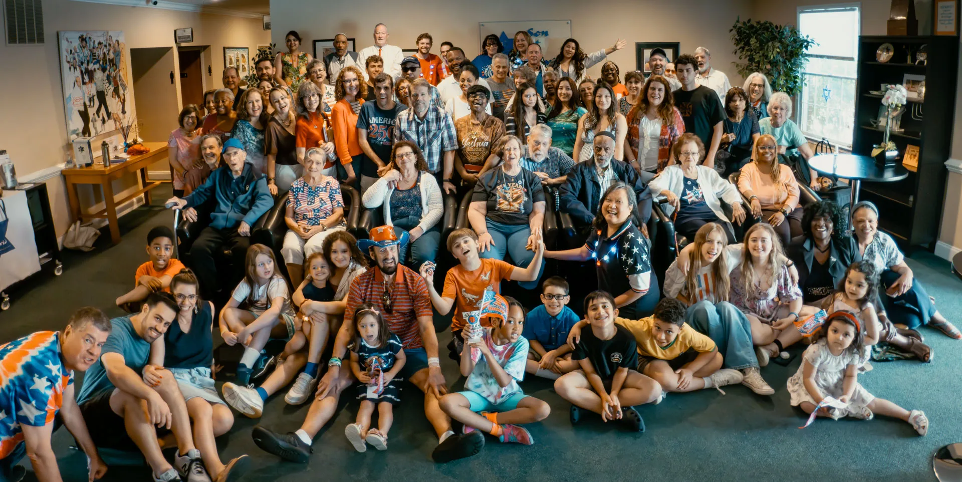 The whole Son of David congregation gathered together for a group photo in the sanctuary — dozens of members of every age, many in orange and red-white-and-blue, seated and standing together with children in the front rows.